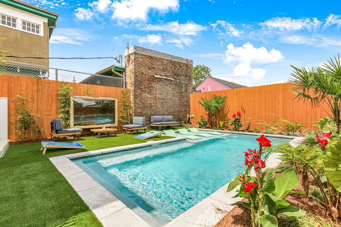 Sunny urban backyard pool oasis with turquoise rectangular pool, lounge seating, cornhole boards, artificial turf and tropical plants with red blooms.
