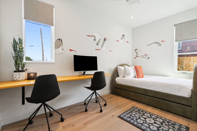Bright modern bedroom with study nook, featuring a long yellow wall-mounted desk with two black swivel chairs and a monitor, potted snake plant, twin daybed with white linens and orange accent pillow, flying bird wall decals, hardwood floor and two windows letting in natural light.