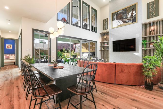 Bright modern open-concept living and dining room with a long dark wood table and black spindle chairs, globe pendant light, rust-orange sectional, wall-mounted TV, tall windows, sliding glass doors to a sunny backyard, hardwood floors and potted plants.