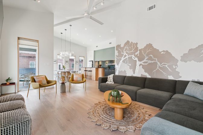 Bright modern urban open-plan living room and kitchen with high ceilings, gray sectional sofa, botanical wall mural, wood coffee table on a woven rug, mustard accent chairs and balcony view.