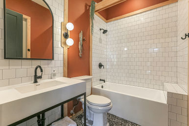 Modern bathroom with white subway tile, terracotta accent wall, arched mirror, matte black fixtures, rectangular vanity sink, toilet, bathtub-shower combo and speckled terrazzo-style floor.