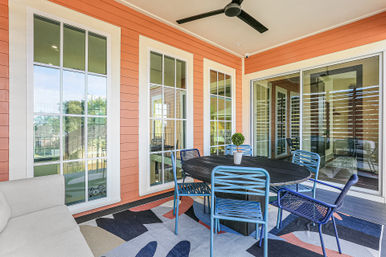 Bright covered patio with coral siding and white-trim windows, round black dining table surrounded by blue metal chairs, small potted topiary on a colorful geometric outdoor rug, sliding glass doors and overhead ceiling fan.