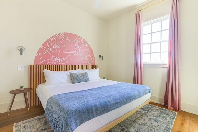 Sunlit modern bedroom with king bed, slatted wood headboard and pink semicircle floral mural, blue throw blanket, pink curtains, small wooden side table with succulent, hardwood floors and patterned area rug.