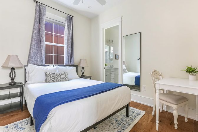 Cozy bright modern bedroom with white bedding and blue throw on a queen bed, patterned accent pillow, dark nightstands with lamps, gray curtains at the window, full-length mirror, white desk and chair with potted plant, hardwood floors and doorway to an en-suite bathroom.