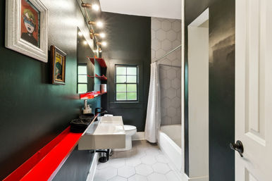 Sleek modern bathroom with dark walls and a pop of red from floating shelves, wall-mounted sink, vanity lights, white hex tile floor and tub with gray hex-tiled surround.