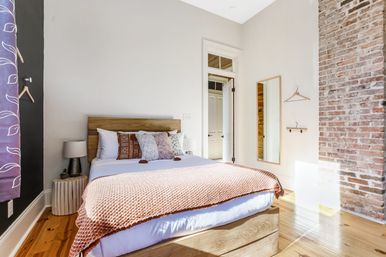 Sunlit cozy urban loft bedroom with wooden platform bed, textured rust throw and patterned pillows, exposed brick accent wall, full-length mirror, bedside lamp and warm hardwood floors.