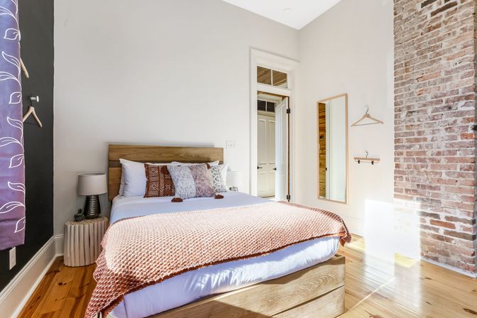 Sunlit cozy urban loft bedroom with wooden platform bed, textured rust throw and patterned pillows, exposed brick accent wall, full-length mirror, bedside lamp and warm hardwood floors.