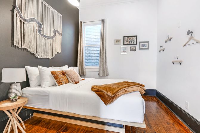 Cozy boho bedroom with macramé wall hanging, white linens and patterned throw pillows on a wooden platform bed, tan blanket, gallery wall and hardwood floors.
