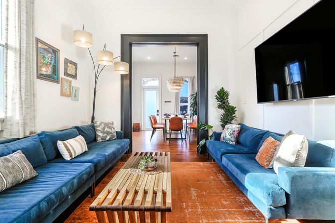 Bright modern living room with twin blue velvet sofas, wooden slatted coffee table on an orange rug, large wall-mounted TV, arc floor lamp and plants, open-concept view into dining area with chandelier.