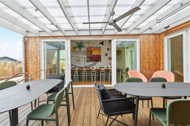 Bright covered wooden deck patio with translucent roof, modern outdoor dining tables and mixed seating (green chairs, black wicker and pink lounge chairs), ceiling fan, and sliding glass doors opening to an indoor dining room with a colorful mural.