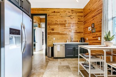 Bright cozy kitchen with knotty wood-paneled walls, stainless-steel refrigerator and dishwasher, gray lower cabinets, farmhouse sink, tiled floor, white metal island with shelves, potted plant by the window, and a decorative Louisiana state outline on the wall.