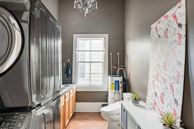 Home laundry and utility room with stacked stainless washer and dryer, wood cabinets and sink under a window, water heater and toilet, leaning flamingo-patterned artwork, terracotta tile floor and small potted plants.