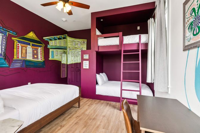 Cozy boutique guest room with deep magenta walls and colorful house mural, large bed on the left and built-in bunk beds with ladder on the right, wood floor, ceiling fan, desk and window with light curtains.