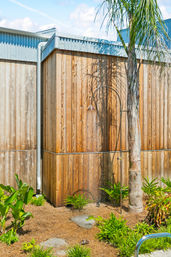 Sunlit poolside outdoor shower on a curved metal pipe by a palm tree against a tall wooden fence in a tropical backyard garden