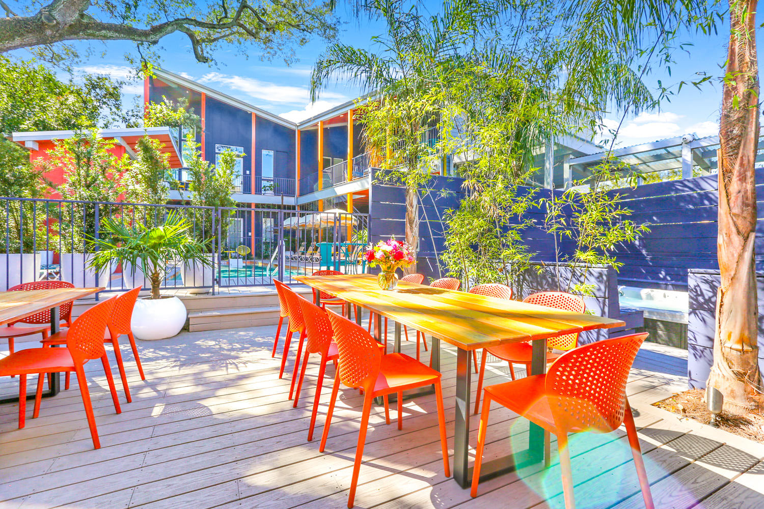 Vibrant sunny outdoor patio with a long wooden dining table and bright orange chairs on a deck beside a pool in a modern courtyard framed by tropical plants and balconies.
