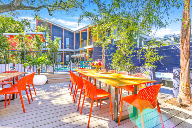 Vibrant sunny outdoor patio with a long wooden dining table and bright orange chairs on a deck beside a pool in a modern courtyard framed by tropical plants and balconies.