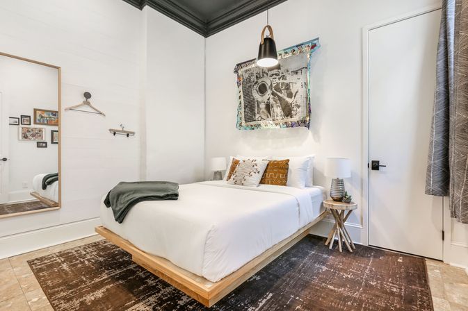 Cozy modern bedroom with floating wooden platform bed, crisp white linens, patterned throw pillows, hanging black pendant light, eclectic wall art, round side table with lamp, full-length mirror and textured area rug.