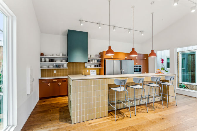 Bright modern open-concept kitchen with tiled breakfast bar, four metal bar stools, orange pendant lights, stainless steel appliances, open shelving and warm hardwood floors
