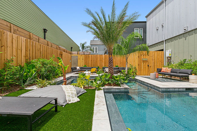 Modern urban backyard with a rectangular blue pool and central palm tree, lush tropical plants, wooden privacy fence, artificial turf and cushioned outdoor lounge seating with colorful accent chairs.