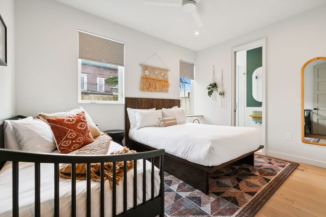 Bright modern bedroom with boho accents: dark wood platform bed with white linens, black spindle daybed with rust-pattern pillow, woven wall hanging, hanging plants, geometric area rug and open ensuite bathroom.
