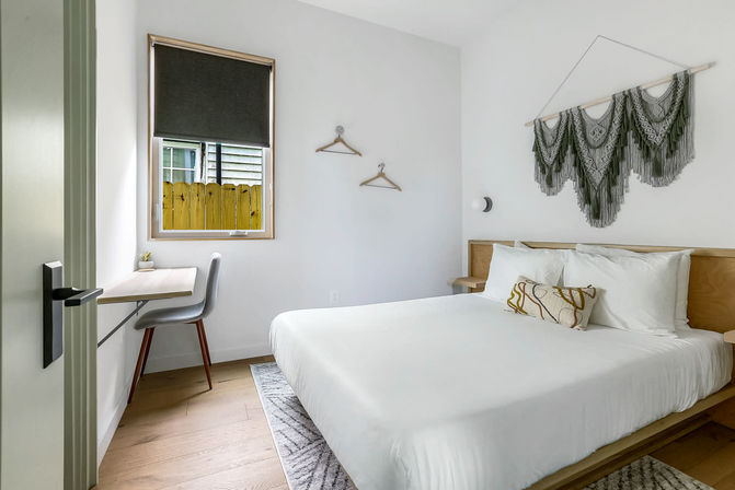 Minimal modern bedroom with white linens on a wooden platform bed, decorative gray macramé wall hanging above the headboard, small window-side desk and gray chair with a roller shade, light wood floors and neutral decor.