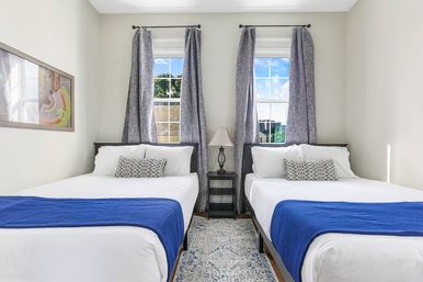 Bright, cozy twin bedroom with two white-made beds, blue throws and patterned pillows, a central black nightstand with lamp, gray curtains framing two windows and a light patterned rug.