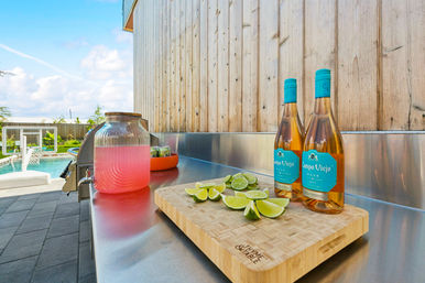 Sunny poolside outdoor kitchen with stainless-steel counter, bamboo cutting board of sliced limes, two rosé wine bottles and a glass dispenser of pink lemonade, wooden fence and pool in background.