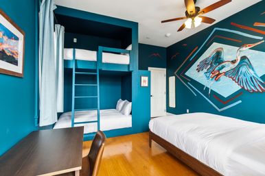 Coastal-inspired teal bedroom with colorful heron mural, built-in bunk beds, queen bed, desk, ceiling fan, and hardwood floor.