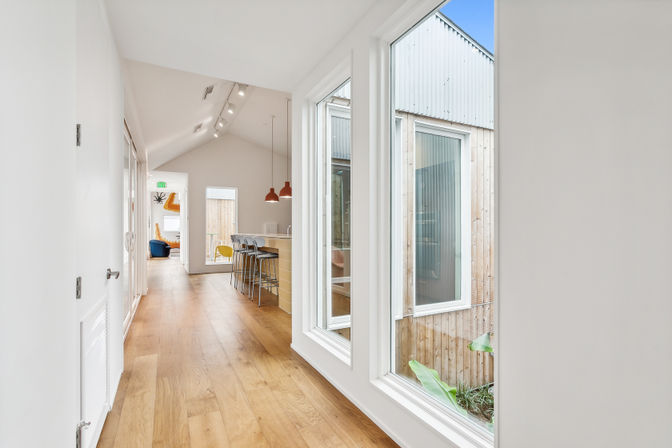 Bright modern hallway with warm hardwood floors, floor-to-ceiling courtyard windows, and an open kitchen island with pendant lights and bar stools.