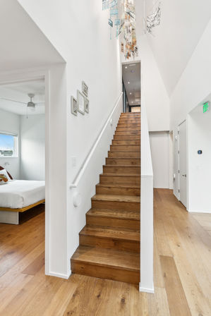 Bright, airy modern interior with warm oak staircase and wide-plank flooring, white walls, high ceiling and hanging mobile artwork; bedroom doorway visible.
