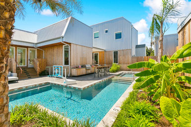 Sunlit coastal modern backyard with lap-style pool and basketball hoop, outdoor kitchen with bar seating, palm and banana plants, and wood-clad contemporary house.