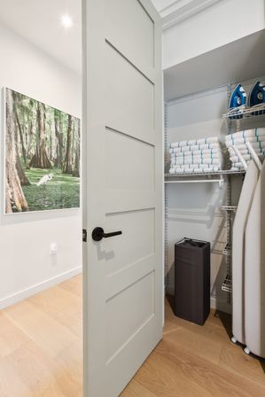 Open paneled interior door revealing a tidy linen closet with neatly folded white towels, blue irons on wire shelves, upright ironing boards and a tall dark laundry hamper against light wood floors and nature wall art.