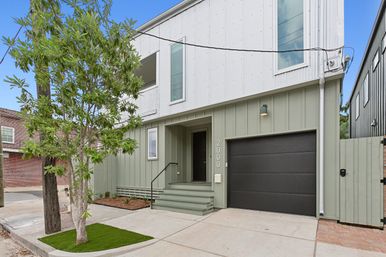 Modern two-story townhouse exterior with sage-green board-and-batten lower siding and white upper cladding, matte black single-car garage door, small front porch with steps, young street tree and concrete driveway — clean urban curb appeal.