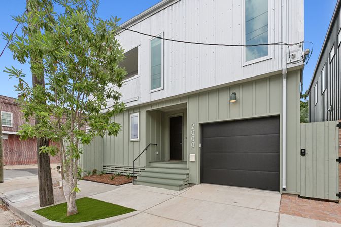 Modern two-story townhouse exterior with sage-green board-and-batten lower siding and white upper cladding, matte black single-car garage door, small front porch with steps, young street tree and concrete driveway — clean urban curb appeal.