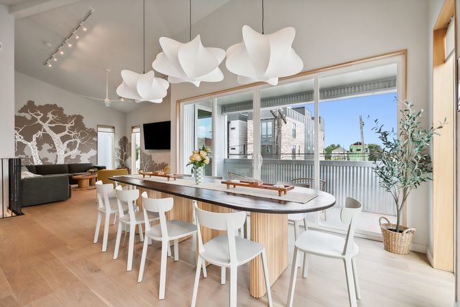 Bright modern open-plan dining and living room in an urban apartment — long wooden dining table with white chairs, sculptural white pendant lights, sliding glass doors to a balcony with city view, tree wall mural and light hardwood floors.