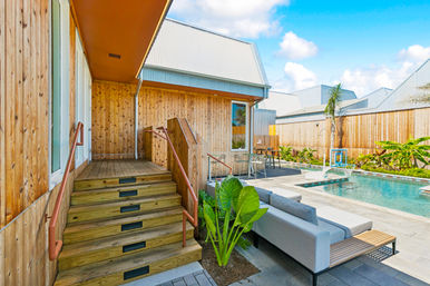 Modern backyard with wooden-clad house and metal roof, steps up to a covered deck, gray outdoor sofa, tropical plants and a rectangular swimming pool on a sunny day