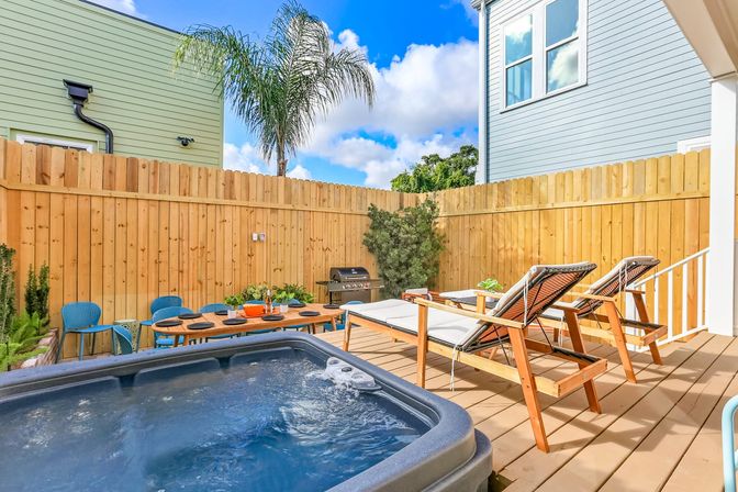 Sunny backyard deck patio with bubbling hot tub, two wooden lounge chairs, outdoor dining table, gas grill, palm tree and tall wooden privacy fence under a bright blue sky.