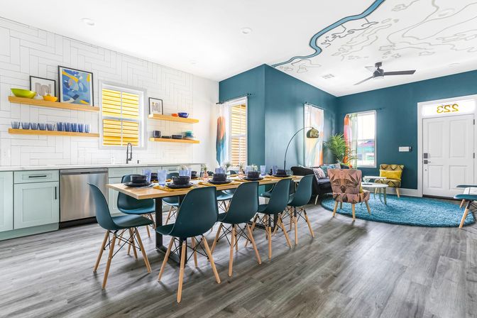 Bright, coastal-inspired open-plan kitchen and living area with teal accent walls, long wooden dining table set for eight with teal chairs, floating wood shelves over white tile backsplash, stainless dishwasher, and a cozy seating area on a turquoise rug beneath a whimsical ceiling mural.