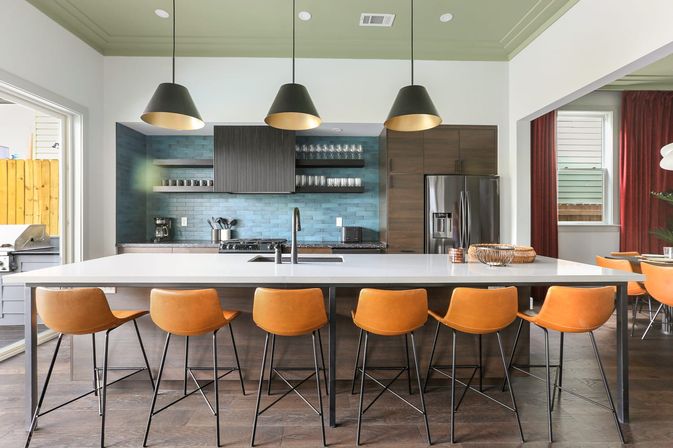 Bright modern kitchen with large white island and breakfast bar, six orange leather stools, three black pendant lights, blue subway tile backsplash, dark wood cabinets and stainless refrigerator.