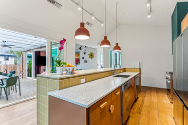 Bright modern open-plan kitchen with speckled island countertop, three terracotta pendant lights, stainless sink and appliances, wood floors, sliding glass doors to a covered outdoor deck with dining area, orchid and fruit bowl on the counter.
