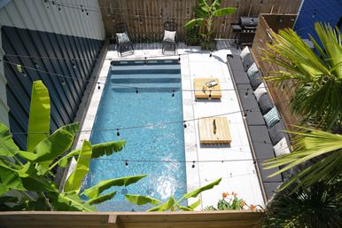 Aerial view of a sunlit backyard rectangular pool and concrete patio with a long gray outdoor sofa, wooden coffee tables, string lights, tropical banana and palm plants, and a barbecue grill.