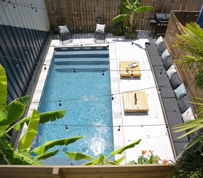 Aerial view of a sunlit backyard rectangular pool and concrete patio with a long gray outdoor sofa, wooden coffee tables, string lights, tropical banana and palm plants, and a barbecue grill.