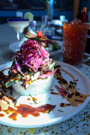 Colorful Asian-inspired rice stack topped with grilled meat, pickled purple cabbage, cucumber strips and nori, drizzled with savory sauces, served on a white plate with a cocktail and tall iced drink on a restaurant table.