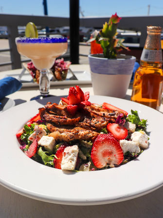 Grilled chicken and fresh strawberry salad with crumbled cheese and mixed greens on a white plate, served al fresco on a sunny patio table with a frozen margarita in a blue-rim glass, a beer bottle and a small potted succulent.