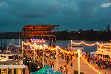 Evening waterfront concert at a marina: outdoor stage under a canopy, warm string lights draped along the dock, crowds gathered on the pier and moored boats on calm water.