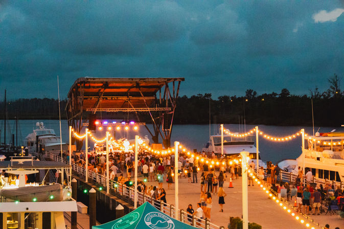Evening waterfront concert at a marina: outdoor stage under a canopy, warm string lights draped along the dock, crowds gathered on the pier and moored boats on calm water.