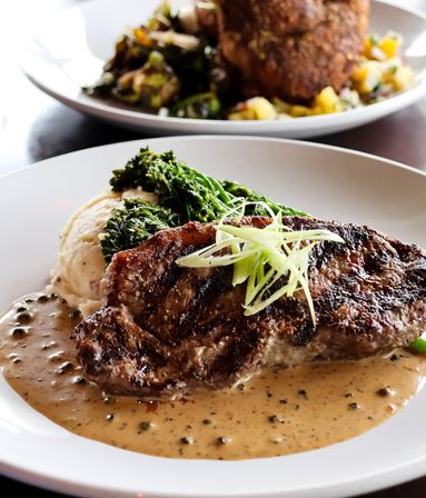 Sizzling grilled steak on a white plate with creamy peppercorn sauce, whipped mashed potatoes, broccolini and thin green onion strips — upscale restaurant entrée.