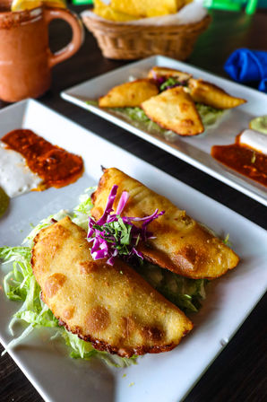 Close-up of Mexican-style crispy fried empanadas on a white square plate over shredded lettuce, topped with purple cabbage and cilantro, served with three dipping sauces and a chips basket on a dark restaurant table.
