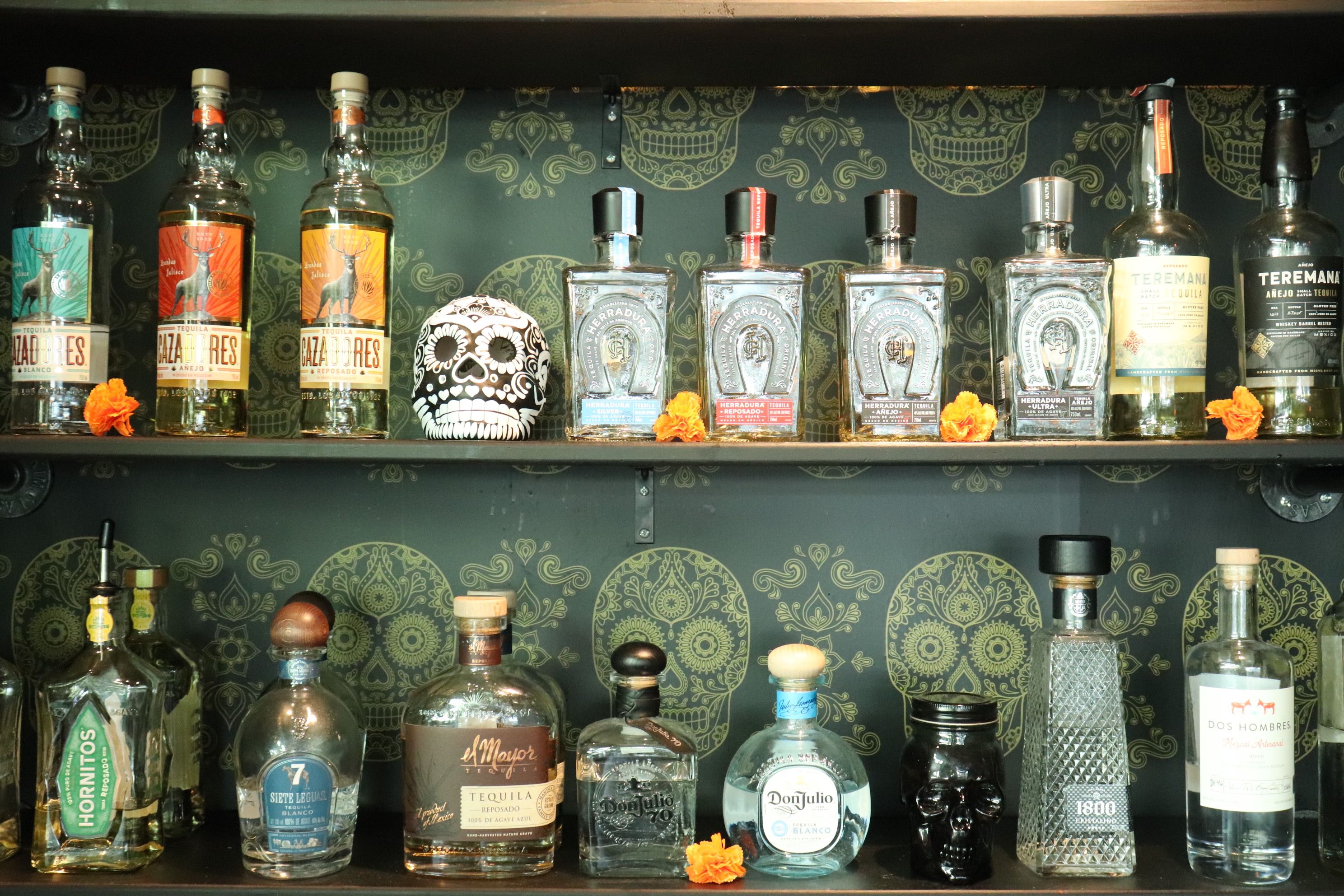 Mexican-themed bar shelf displaying tequila bottles, a decorative Day of the Dead skull, marigold flowers, and green skull-patterned wallpaper.