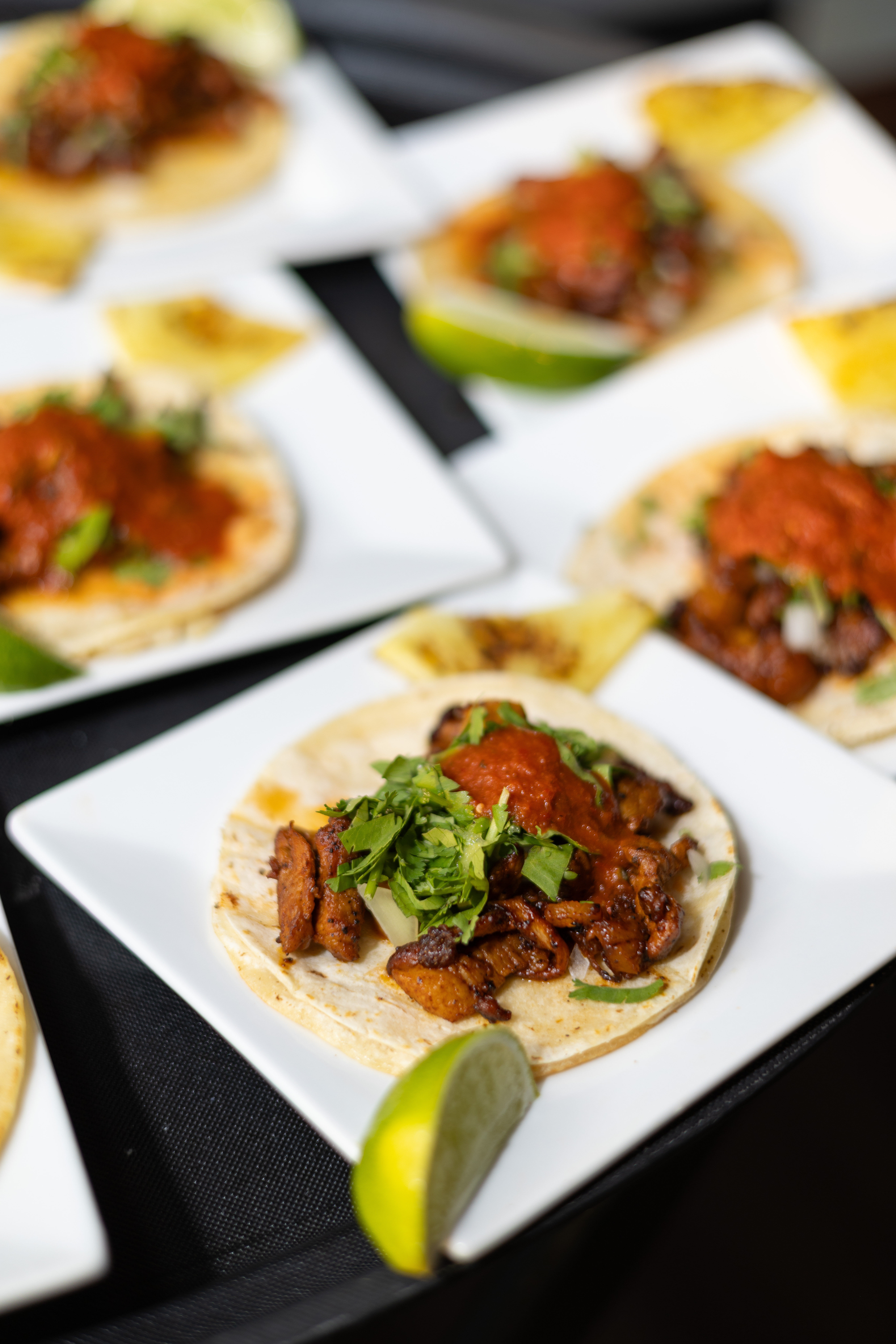 Close-up of Mexican street tacos on corn tortillas with grilled seasoned meat, chopped cilantro and red salsa, served with lime wedges on white plates